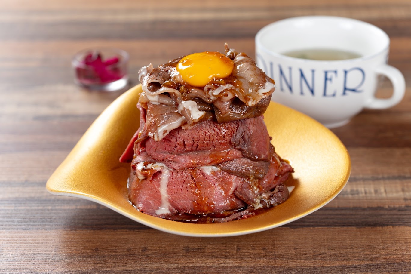 ข้าวหน้าเนื้อสไตล์ญี่ปุ่นวางซ้อนชิ้นหนานุ่ม ราดซอสและไข่แดงดิบ เสิร์ฟบนจานสีทอง บรรยากาศอบอุ่น เหมาะสำหรับสายกินที่วางแผน เที่ยวญี่ปุ่น