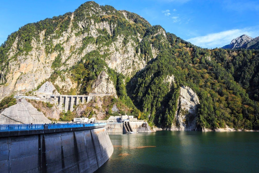 "ภาพถ่ายเขื่อนคุโรเบะ (Kurobe Dam) อันเป็นส่วนหนึ่งของเส้นทางเจแปนแอลป์ ซึ่งเป็นจุดแวะพักยอดนิยมสำหรับการ ขับรถเที่ยว japan alps ด้วยรถเช่าญี่ปุ่น แสดงให้เห็นความยิ่งใหญ่ของสันเขื่อนคอนกรีต โอบล้อมด้วยอ่างเก็บน้ำสีเขียวมรกตและเทือกเขาสูงชันที่ปกคลุมด้วยป่าไม้เปลี่ยนสีในช่วงฤดูใบไม้ร่วง ภายใต้ท้องฟ้าสดใส"
