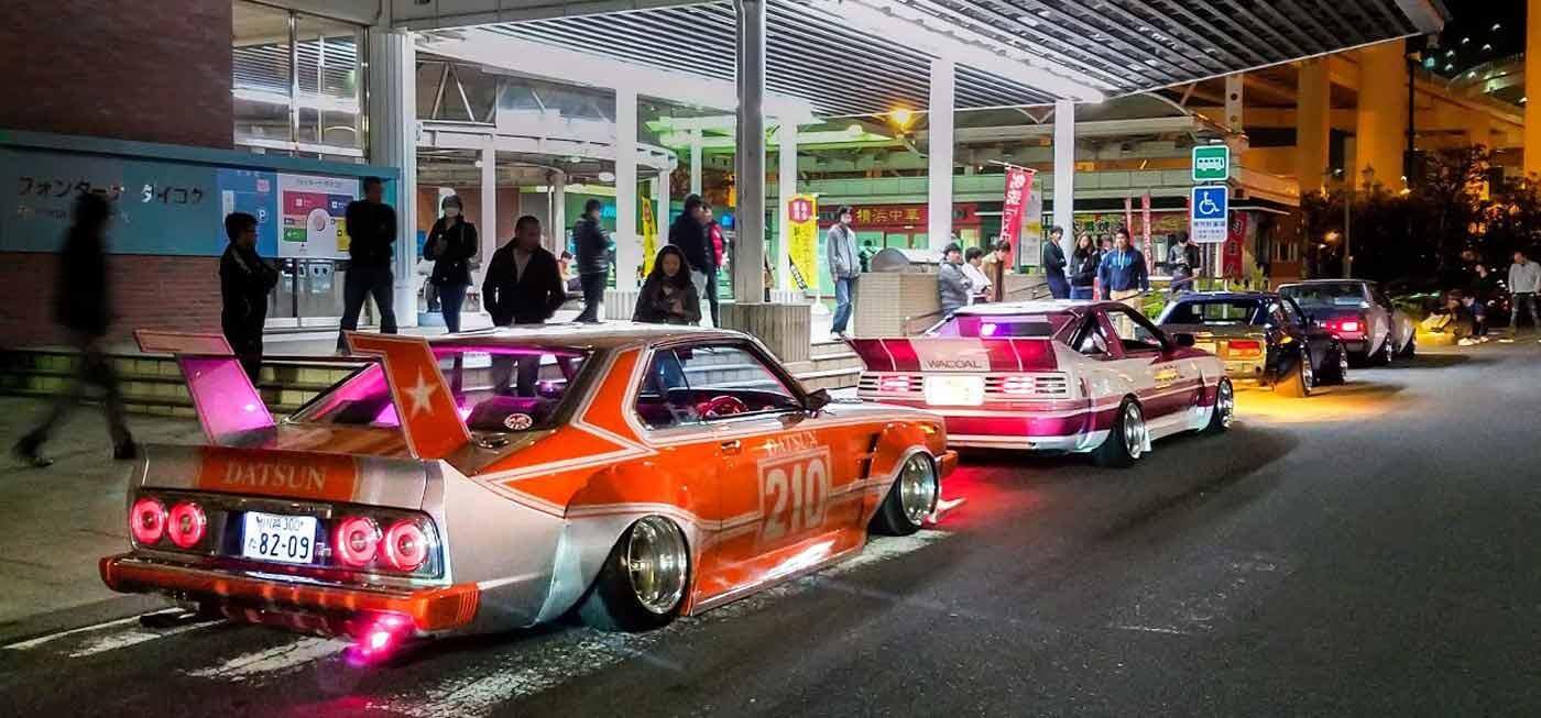 รูปภาพรถคลาสสิก Datsun สไตล์แต่งรถญี่ปุ่น JDM แบบ Kaido Racer / Bosozoku ที่ปรับแต่งอย่างสุดขั้ว พร้อมซูเปอร์สปอยเลอร์ขนาดใหญ่และนีออนนีออนสีชมพู จอดเรียงรายอยู่ที่สถานีบริการ Daikoku PA ในญี่ปุ่นยามค่ำคืน
