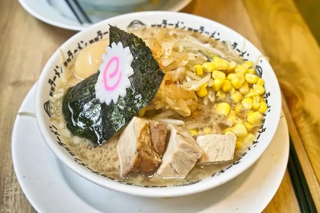 tsukemen กับ ramen ต่างกันยังไง ภาพราเมนญี่ปุ่นเสิร์ฟในชามน้ำซุปร้อน มีหมูชาชู ไข่ต้ม ข้าวโพด ถั่วงอก สาหร่าย และนารูโตะ บนโต๊ะไม้ในร้านอาหารญี่ปุ่น