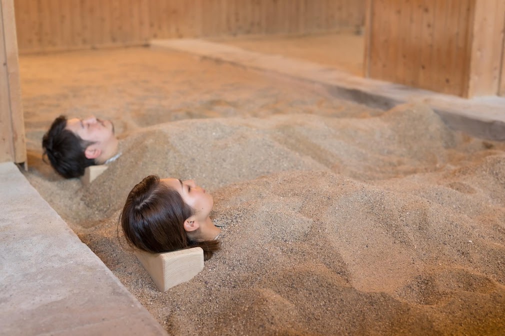 "ชายหญิงนอนผ่อนคลายในบ่อทรายร้อน (Sunayu) ในร่มที่ Hyotan Onsen เบปปุ โดยใช้หมอนไม้รองศีรษะท่ามกลางบรรยากาศการตกแต่งด้วยไม้ที่อบอุ่น"