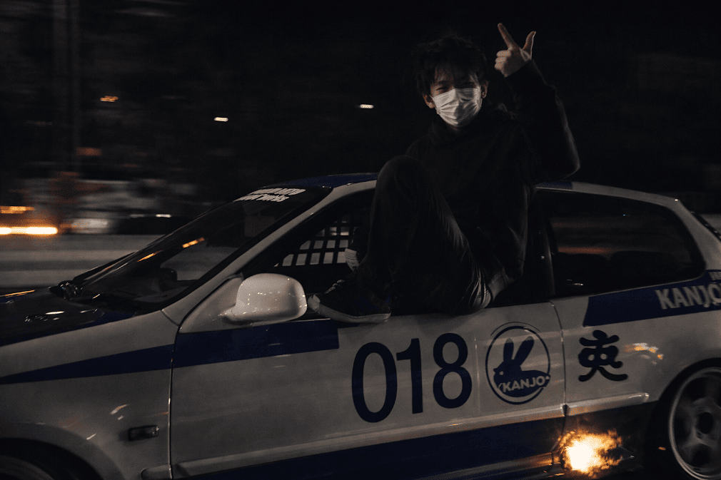 kanjozoku คืออะไร มีที่มาอย่างไร — ภาพชายสวมหน้ากากนั่งบนรถแต่งสไตล์ฮอนด้า Civic หมายเลข 018 ขับบนทางด่วนญี่ปุ่นยามค่ำคืน มีประกายไฟจากท่อไอเสีย สื่อถึงวัฒนธรรมแก๊งรถซิ่งคันโจโซกุที่เกิดจากการแข่งบนทางด่วนวงแหวนโอซาก้า