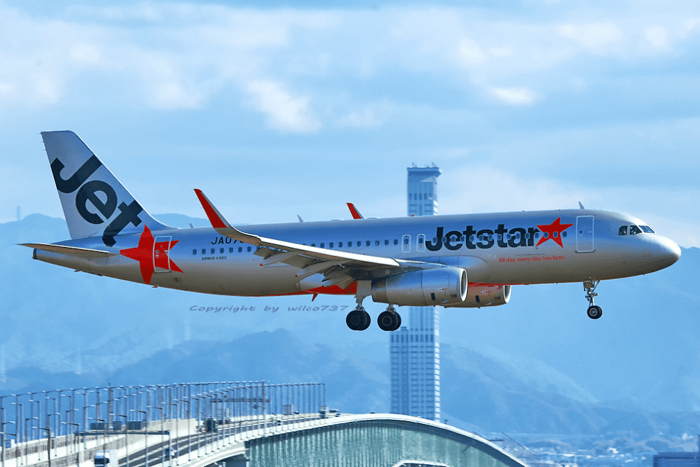 สายการบินของญี่ปุ่น Jetstar Japan เครื่องบิน Airbus A320 กำลังลงจอดที่สนามบิน โดยมีภูเขาและอาคารในเมืองญี่ปุ่นเป็นฉากหลัง แสดงถึงสายการบินต้นทุนต่ำยอดนิยม ✈️🇯🇵