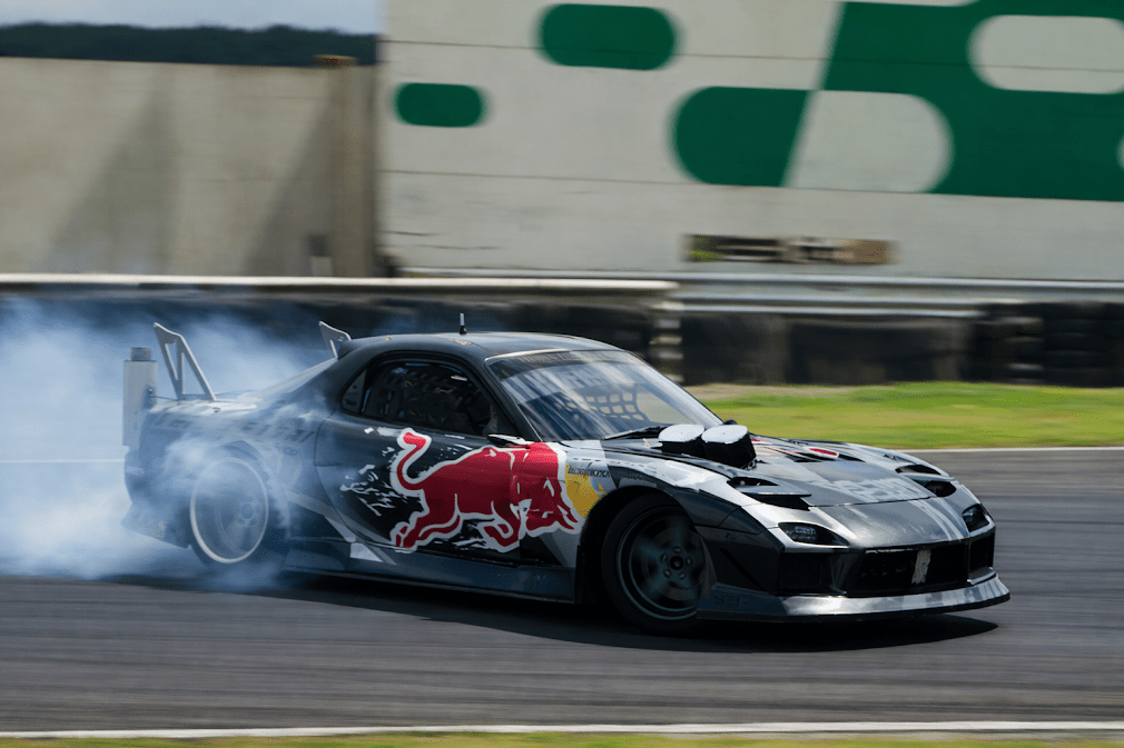 รถ drift ญี่ปุ่น Mazda RX-7 แต่งเต็มกำลังดริฟต์ในสนามแข่ง มีควันยางลอยด้านหลัง พร้อมชุดแต่งสไตล์รถแข่งและลวดลายสปอร์ต แสดงถึงสมรรถนะและเอกลักษณ์ของรถดริฟต์ญี่ปุ่น 🚗💨🇯🇵