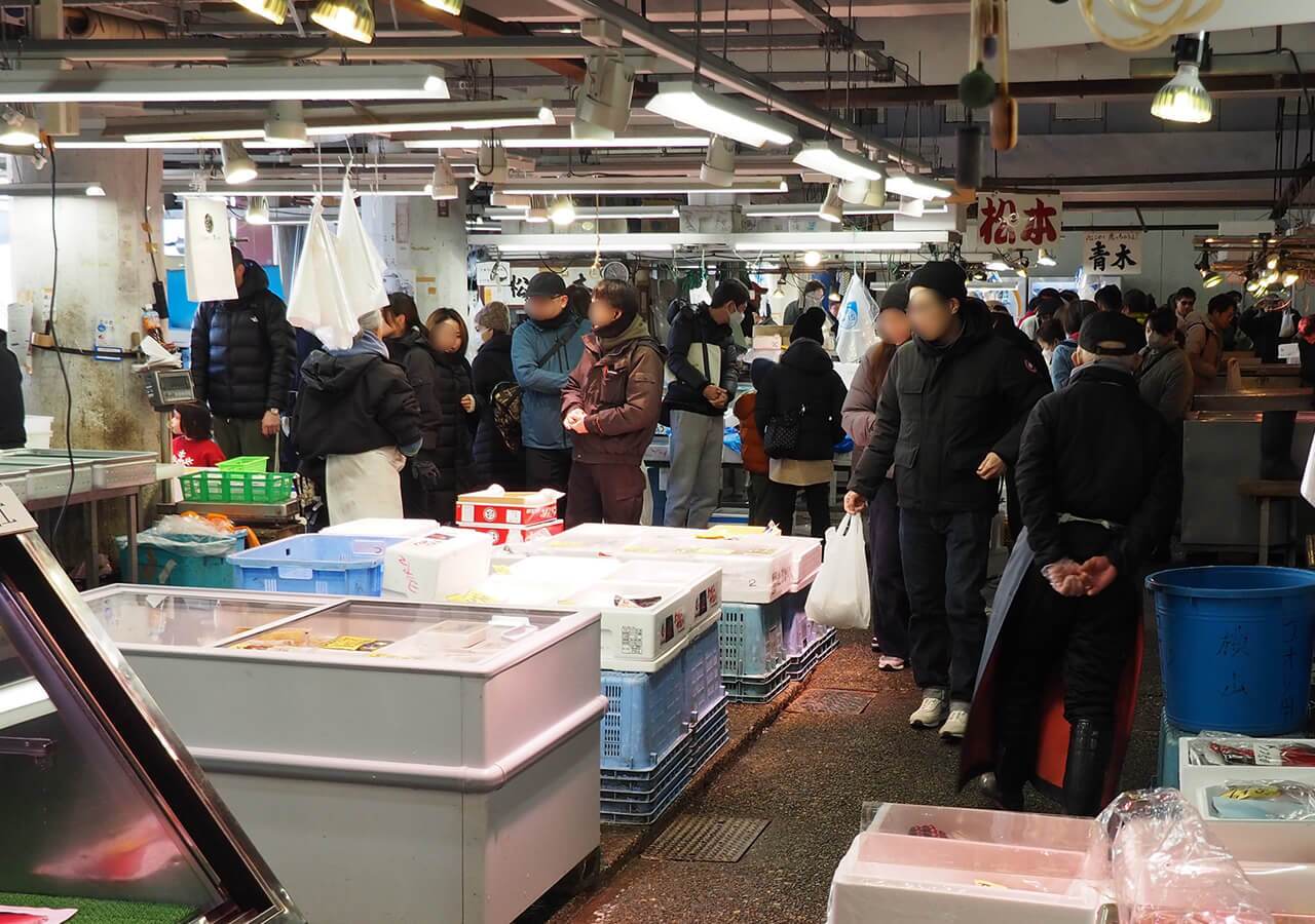 บรรยากาศทางเดินภายใน ตลาดปลา ใน ญี่ปุ่น ที่เนืองแน่นไปด้วยผู้คนและนักท่องเที่ยวในชุดเสื้อกันหนาวหนาๆ กำลังเดินเลือกซื้ออาหารทะเล รอบข้างเต็มไปด้วยแผงค้าที่มีกล่องโฟมและลังพลาสติกใส่ของสดวางเรียงราย พร้อมตู้แช่แข็งกระจกใส ด้านบนเพดานเปิดไฟนีออนสว่างไสว มีป้ายชื่อร้านภาษาญี่ปุ่นแขวนอยู่ และถุงพลาสติกแขวนเตรียมไว้สำหรับบริการลูกค้า ให้ความรู้สึกถึงวิถีชีวิตในตลาดท้องถิ่นที่คึกคัก