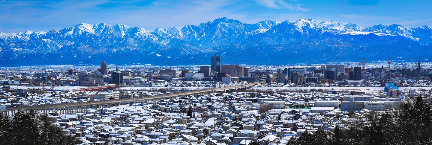 "ภาพถ่ายทัศนียภาพอันกว้างใหญ่ของเทือกเขาแอลป์ญี่ปุ่น (Japan Alps) ที่ปกคลุมด้วยหิมะ โอบล้อมเมืองทาเตยามะและแม่น้ำที่แข็งตัวในช่วงฤดูหนาว แสดงถึงความสวยงามของเส้นทางยอดนิยมสำหรับ ขับรถเที่ยว japan alps ด้วยรถเช่าญี่ปุ่น ภายใต้ท้องฟ้าสีฟ้าคราม"