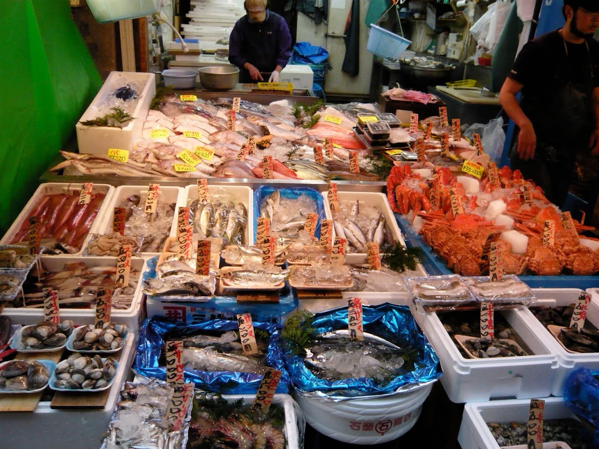 บรรยากาศแผงขายอาหารทะเลสดหลากชนิดใน ตลาดปลา ใน ญี่ปุ่น (ตลาดสึคิจิ) ที่จัดวางสินค้าในกระบะโฟมและถาดแช่น้ำแข็งอย่างหนาแน่น ด้านหน้ามีหอยแครง หอยแมลงภู่ กุ้งสด และปลาตัวเล็ก ถัดไปเป็นปลาหมึกสด ปลากระบอก และโซนปูยักษ์ฮอกไกโดสีส้มสะดุดตา ทุกถาดมีป้ายราคากระดาษเขียนด้วยพู่กันญี่ปุ่นปักอยู่ โดยมีพ่อค้ากำลังยืนเตรียมสินค้าอยู่ด้านหลังร้าน
