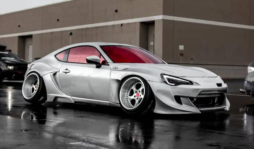 รถสปอร์ตแต่ง สไตล์แต่งรถญี่ปุ่น JDM สีเงินสุดเท่ พร้อมชุดแต่ง Wide Body รอบคันและกระจกสีชมพูสะท้อนแสง โดดเด่นบนพื้นถนนเปียกน้ำ