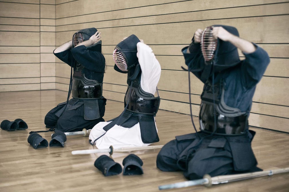 Three Kendo practitioners in a Japanese dojo, sitting in seiza and carefully adjusting their men (masks). This image captures a moment of ritualized preparation, showing part of the bogu and the process of gearing up, which helps answer ฟันดาบ kendo คืออะไร by demonstrating its emphasis on discipline and protective equipment. Shinai (bamboo swords) and kote (gloves) are visible on the polished floor. The scene is set against a traditional slatted wooden wall.