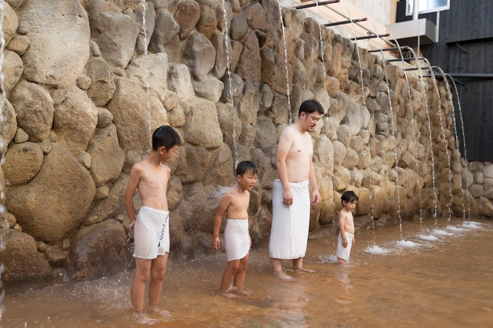 "ครอบครัวอาบน้ำใต้สายน้ำตก Takiyu ที่ Hyotan Onsen เบปปุ โดยมีผนังหินก้อนใหญ่เป็นฉากหลัง"