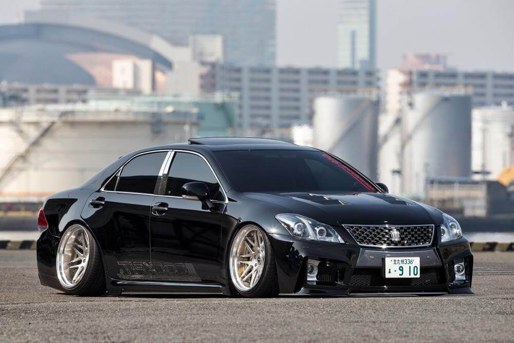 รถซีดานหรู Toyota Crown สีดำเงา ตกแต่งในสไตล์ VIP Style ที่สะท้อนกลิ่นอาย สไตล์แต่งรถญี่ปุ่น JDM ด้วยการลดความสูงของตัวรถ (Stanced) ใส่ล้อแม็กโครเมียมลายซี่ถี่ขนาดใหญ่หน้าตื้นหลังลึก และชุดแต่งรอบคัน ในฉากหลังที่เป็นพื้นที่ท่าเรืออุตสาหกรรม