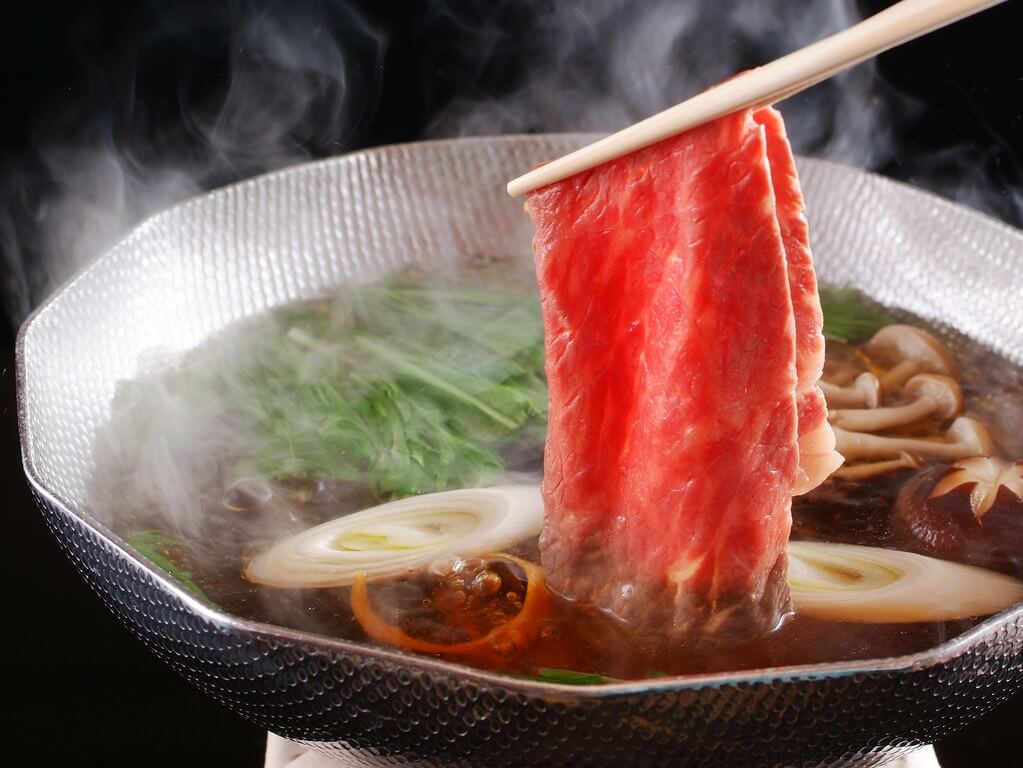 ภาพชาบูสไตล์ญี่ปุ่นอธิบาย shabu คืออะไร ด้วยการคีบเนื้อวัวสไลซ์บางลงลวกในหม้อน้ำซุปร้อน มีไอน้ำลอยขึ้นพร้อมผักสด เห็ด และต้นหอม แสดงเอกลักษณ์ของชาบูที่เน้นความสดของวัตถุดิบและการลวกเนื้อในน้ำซุป"