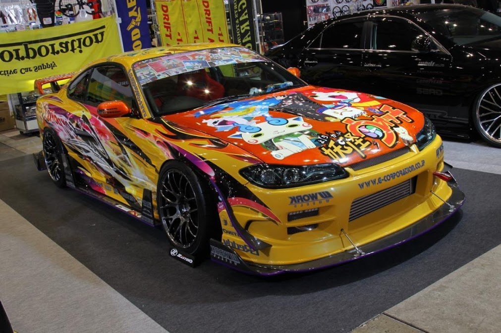 รถ Nissan Silvia S15 สีเหลืองส้มที่ตกแต่งในสไตล์ "Itasha" ด้วยลายการ์ตูนอนิเมะ "Yo-kai Watch" ทั่วทั้งคัน โดดเด่นเป็นสง่าในงานนิทรรศการที่รวม สไตล์แต่งรถญี่ปุ่น JDM รูปแบบต่างๆ ไว้ด้วยกัน ซึ่งเป็นตัวอย่างที่ชัดเจนของวัฒนธรรมการแต่งรถที่เป็นเอกลักษณ์และได้รับความนิยมอย่างสูงในกลุ่มคนรักรถสไตล์ JDM