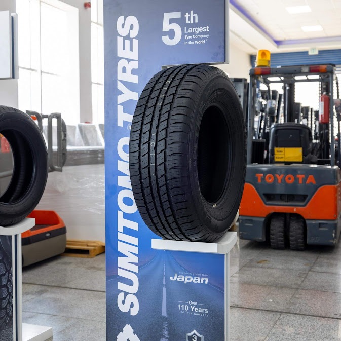ยางรถยนต์ Sumitomo บนแท่นโชว์สินค้าสีน้ำเงินที่ระบุว่าเป็นยี่ห้อยางญี่ปุ่นยอดนิยม และเป็นบริษัทผลิตยางที่ใหญ่เป็นอันดับ 5 ของโลก พร้อมประสบการณ์ยาวนานกว่า 110 ปี โดยมีฉากหลังเป็นโกดังสินค้าและรถยก Toyota
