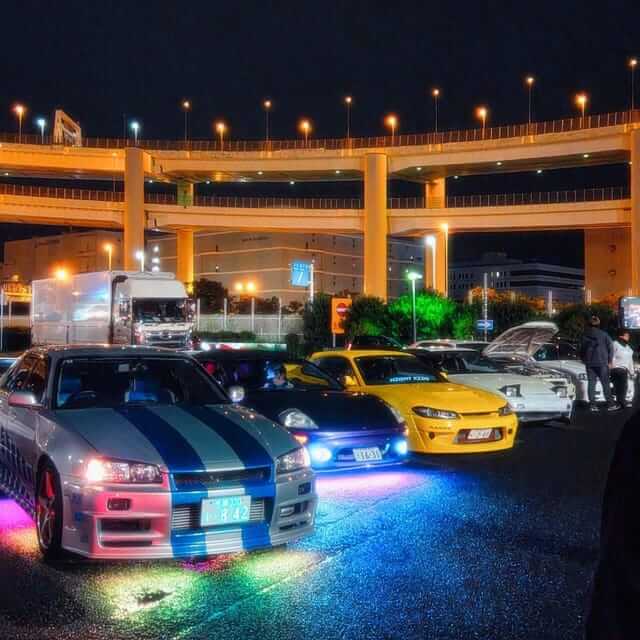 บรรยากาศยามค่ำคืนที่ จุดรวมตัวรถซิ่ง ใน Tokyo เผยให้เห็นกลุ่มรถสปอร์ตญี่ปุ่นแต่งเต็มระบบจอดเรียงราย ทั้ง Nissan Skyline สีเงินคาดฟ้า และ Nissan Silvia S15 สีเหลืองที่ส่องแสงไฟสีสันสดใส ท่ามกลางบรรยากาศคึกคักและแสงไฟจากทางยกระดับ