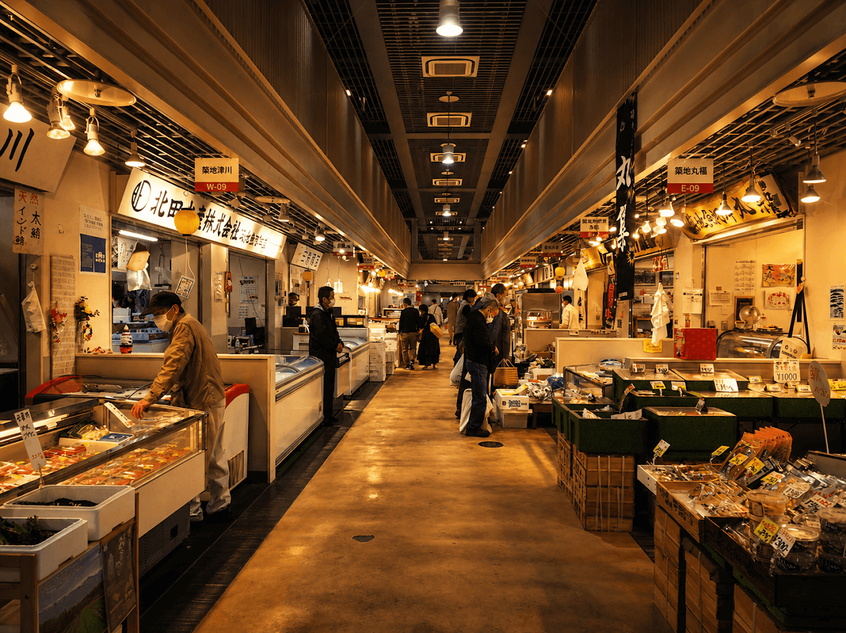 บรรยากาศทางเดินภายในอาคารค้าปลีกที่สะอาดและทันสมัยของ ตลาดปลา ใน ญี่ปุ่น ขนาบข้างด้วยร้านค้าและแผงขายอาหารทะเลแช่เย็นที่เป็นระเบียบ ตกแต่งด้วยแสงไฟสีส้มอบอุ่นสไตล์วอร์มไวท์ ด้านบนมีป้ายร้านอักษรคันจิและป้ายรหัสตำแหน่งสีส้มเด่นชัด มีพ่อค้าแม่ค้าสวมหน้ากากอนามัยกำลังจัดเตรียมสินค้าในตู้แช่กระจกใส และมีลูกค้าเดินเลือกซื้อของอยู่ตรงกลางทางเดิน สะท้อนถึงการผสมผสานวิถีตลาดดั้งเดิมเข้ากับความสะอาดและเป็นสัดส่วนสะดุดตา