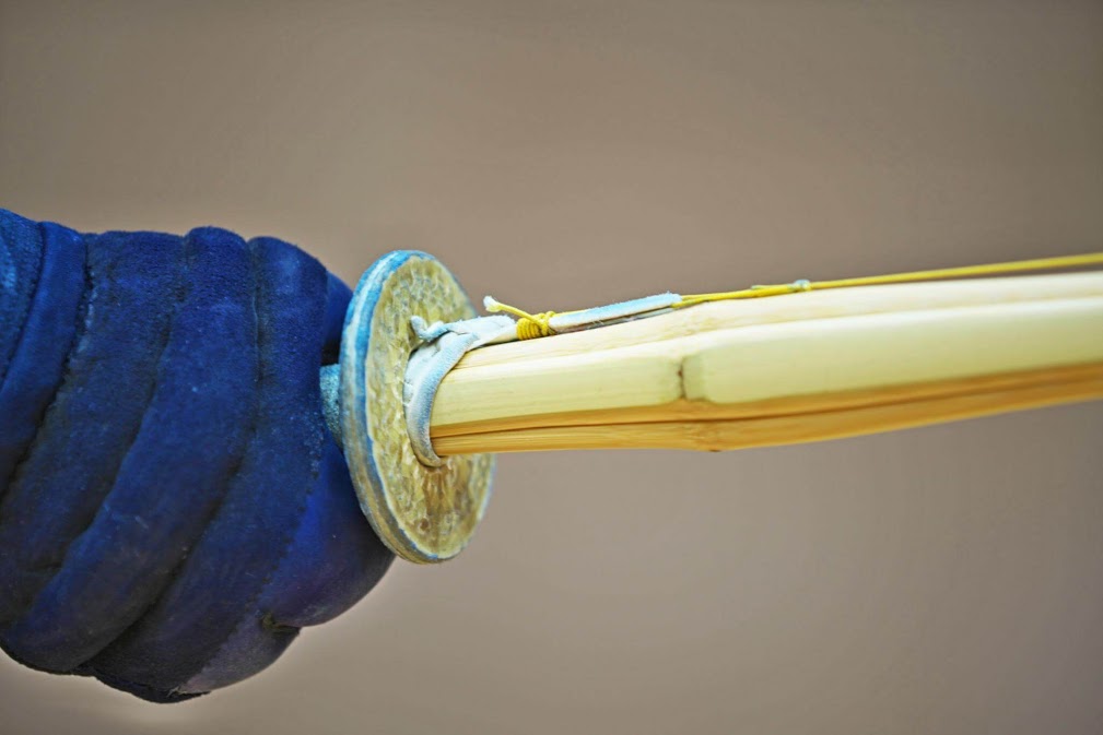 A detailed close-up photograph focusing on the hilt section of a Kendo bamboo sword (shinai). A gloved hand, covered in a ribbed blue padding common to Kendo kote, grips the light-colored bamboo hilt (tsuka). A distinctive, patterned gold-colored metal handguard (tsuba) separates the hilt from the blade. White fabric/leather bindings and a yellow-green cord are visible near the hilt, and the light wood grain of the shinai blade extends to the right against a blurred background. This close-up view of the essential Kendo training tools provides a visual component to understanding ฟันดาบ kendo คืออะไร.
