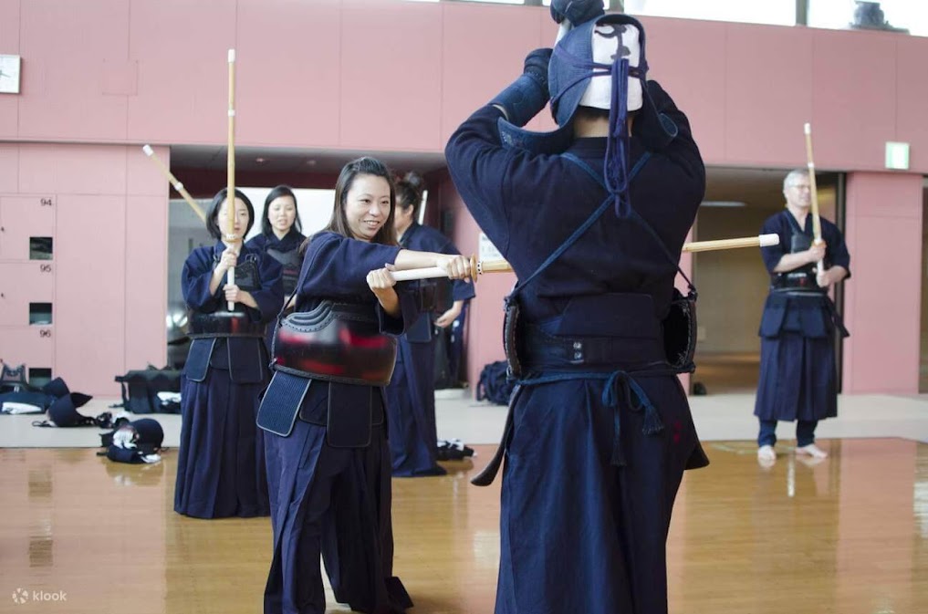 ภาพถ่ายภายในโรงฝึก Kendo (dojo) แบบญี่ปุ่น แสดงผู้คนกำลังฝึกซ้อมศิลปะการต่อสู้แบบเคนโด ผู้หญิงเอเชียที่ยิ้มสวมชุดป้องกัน bogu และถือ shinai (ดาบไม้) กำลังฝึกท่ากับชายที่สวม men (หน้ากาก) และ bogu เต็มชุด ภาพนี้ช่วยอธิบายว่า ฟันดาบ kendo คืออะไร โดยแสดงบรรยากาศการเรียนรู้และการฝึกใช้อุปกรณ์ มีผู้เข้าร่วมคนอื่นๆ ใน bogu ยืนอยู่เบื้องหลัง มีโลโก้ klook ที่มุมซ้ายล่าง