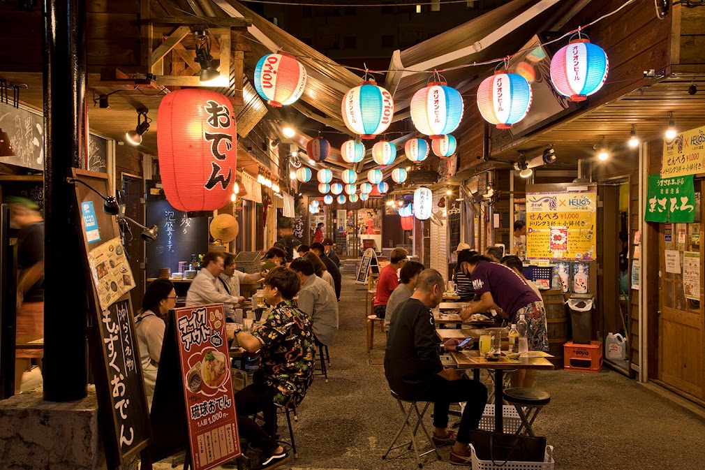 ร้านอาหารสไตล์อิซากายะในโอกินาว่าช่วงกลางคืน ตกแต่งด้วยโคมไฟญี่ปุ่นหลากสี แขวนเรียงรายเหนือโต๊ะที่มีผู้คนนั่งรับประทานอาหารอย่างคึกคัก สะท้อนบรรยากาศท้องถิ่นที่มีชีวิตชีวา เหมาะสำหรับนักท่องเที่ยวที่เดินทางด้วย รถเช่าญี่ปุ่น โอกินาว่า เพื่อสัมผัสวัฒนธรรมการกินดื่มแบบญี่ปุ่นแท้ ๆ