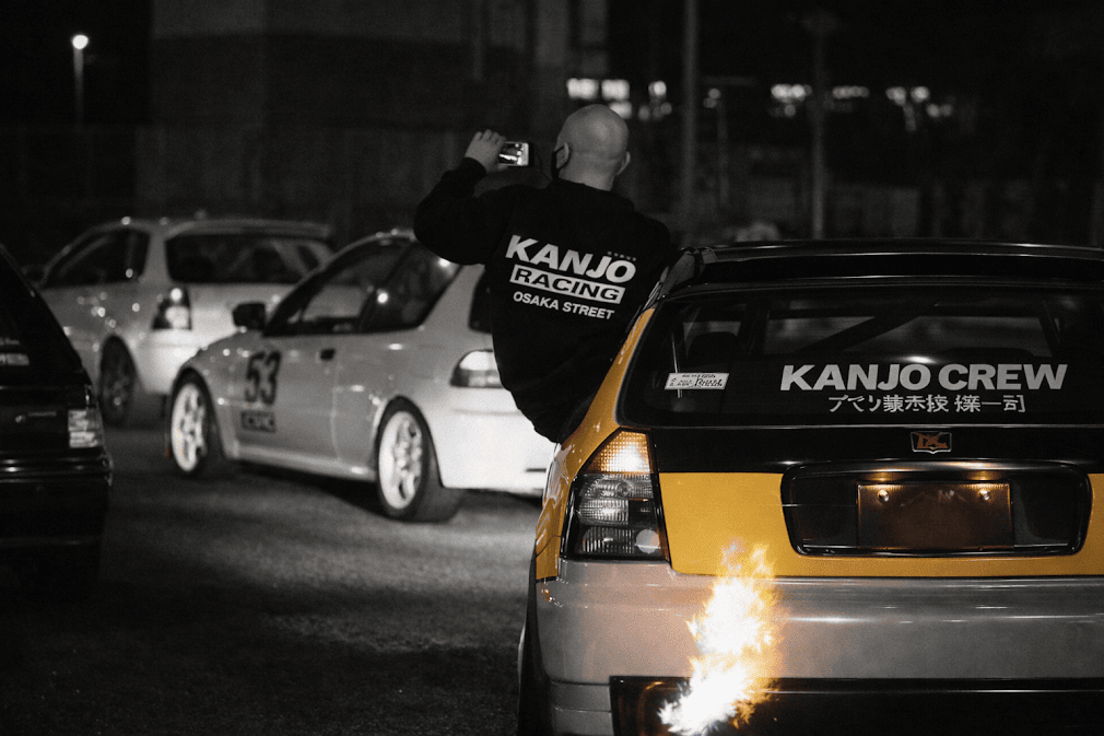 kanjozoku คืออะไร มีที่มาอย่างไร — ภาพกลุ่มรถแต่งสไตล์ Honda Civic จอดรวมตัวบนถนนโอซาก้ายามค่ำคืน ชายสวมเสื้อ Kanjo Racing ถ่ายภาพ มีสติ๊กเกอร์ Kanjo Crew และเปลวไฟจากท่อไอเสีย สะท้อนวัฒนธรรมแก๊งรถซิ่งคันโจโซกุที่กำเนิดจากทางด่วนวงแหวนโอซาก้า