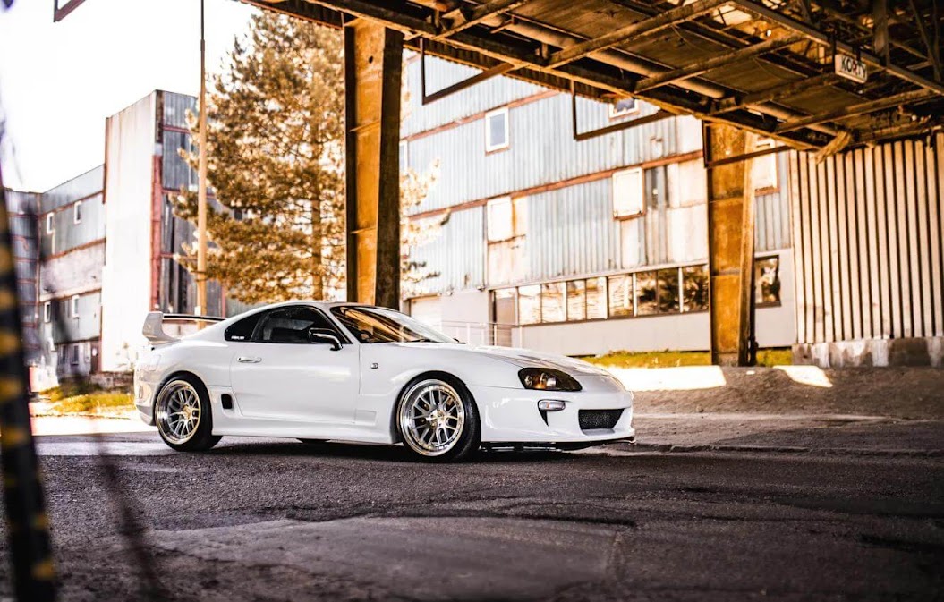 Toyota Supra vs Nissan Skyline comparison featuring a white Toyota Supra Mk4 parked under an industrial structure, showcasing its iconic sporty design, wide stance, and performance styling in an urban setting