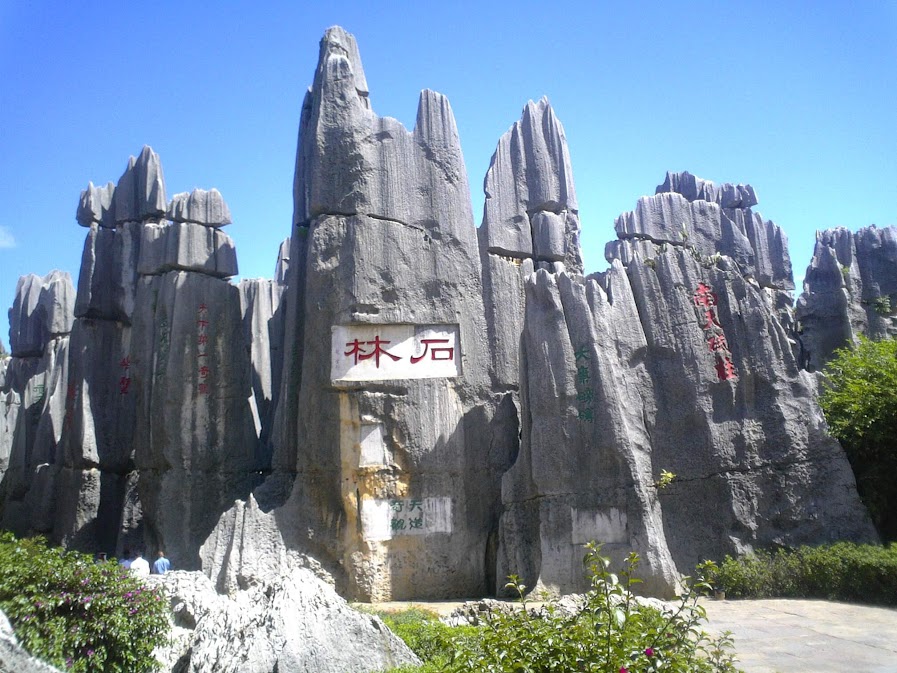 ภาพกลุ่มแท่งหินปูนสูงตระหง่านรูปร่างแปลกตาของอุทยานป่าหินสือหลิน (Shilin Stone Forest) ซึ่งเป็น ที่เที่ยว คุนหมิง ที่มีชื่อเสียง โดดเด่นด้วยการสลักอักษรจีนสีแดงบนหน้าผาหิน ตัดกับท้องฟ้าสีครามสดใสและมีพุ่มไม้สีเขียวประดับอยู่บริเวณฐานล่าง