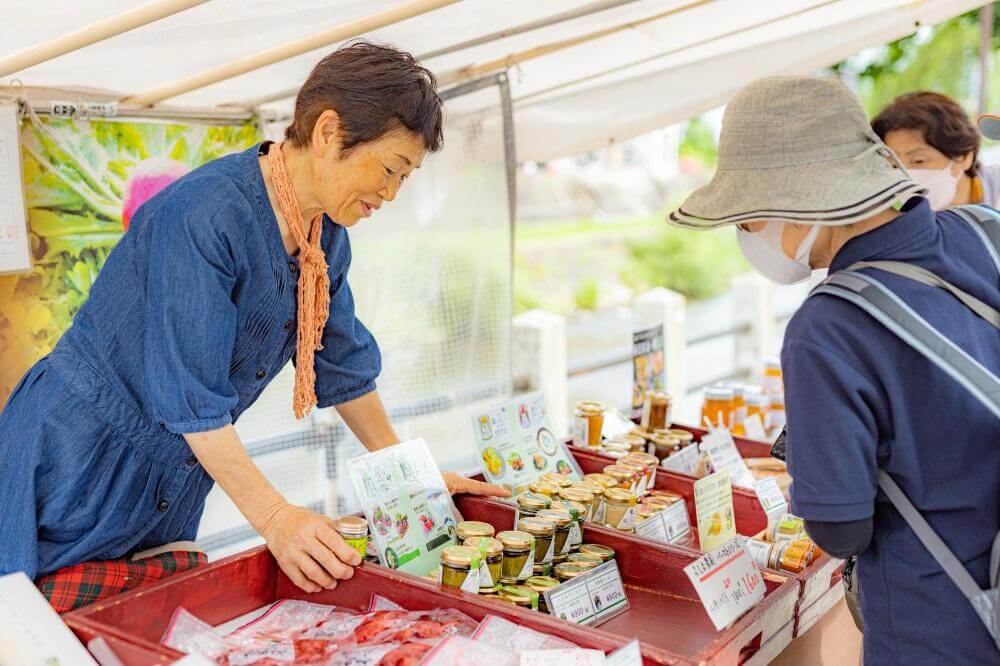 "บรรยากาศตลาดเช้าในท้องถิ่นที่นักท่องเที่ยวสามารถแวะซื้อของกินระหว่างขับรถเที่ยว japan alps ด้วยรถเช่าญี่ปุ่น โดยมีคุณป้าชาวญี่ปุ่นกำลังแนะนำสินค้าจากฟาร์มอย่างเป็นกันเอง"
