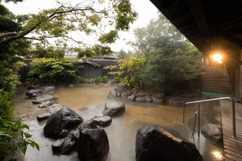 บ่อน้ำพุร้อนกลางแจ้ง (rotenburo) หินธรรมชาติที่ Hyotan Onsen Beppu ล้อมรอบด้วยพุ่มไม้และต้นไม้เขียวขจี พร้อมบันไดไม้และโคมไฟที่ส่องแสงอบอุ่นในตอนเย็น"