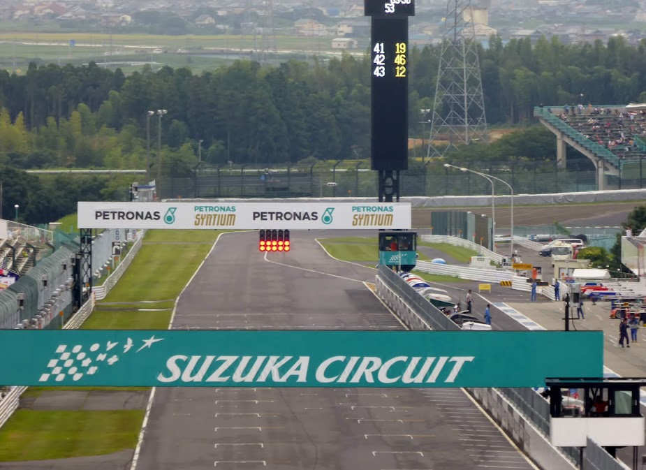 ภาพทางตรงเส้นสตาร์ทของ Suzuka Circuit ใน ญี่ปุ่น มีไฟสตาร์ทสีแดงเหนือแทร็ก ป้าย PETRONAS และอัฒจันทร์ผู้ชมด้านข้าง เหมาะกับคำค้นหา “สนามแข่งรถญี่ปุ่น มีที่ไหนบ้าง”
