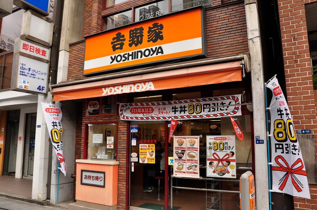 หน้าร้าน Yoshinoya (โยชิโนยะ) ร้านข้าวหน้าเนื้อที่ญี่ปุ่น พร้อมป้ายโปรโมชั่น "80 เยน ส่วนลด" และบรรยากาศริมถนนในโตเกียว