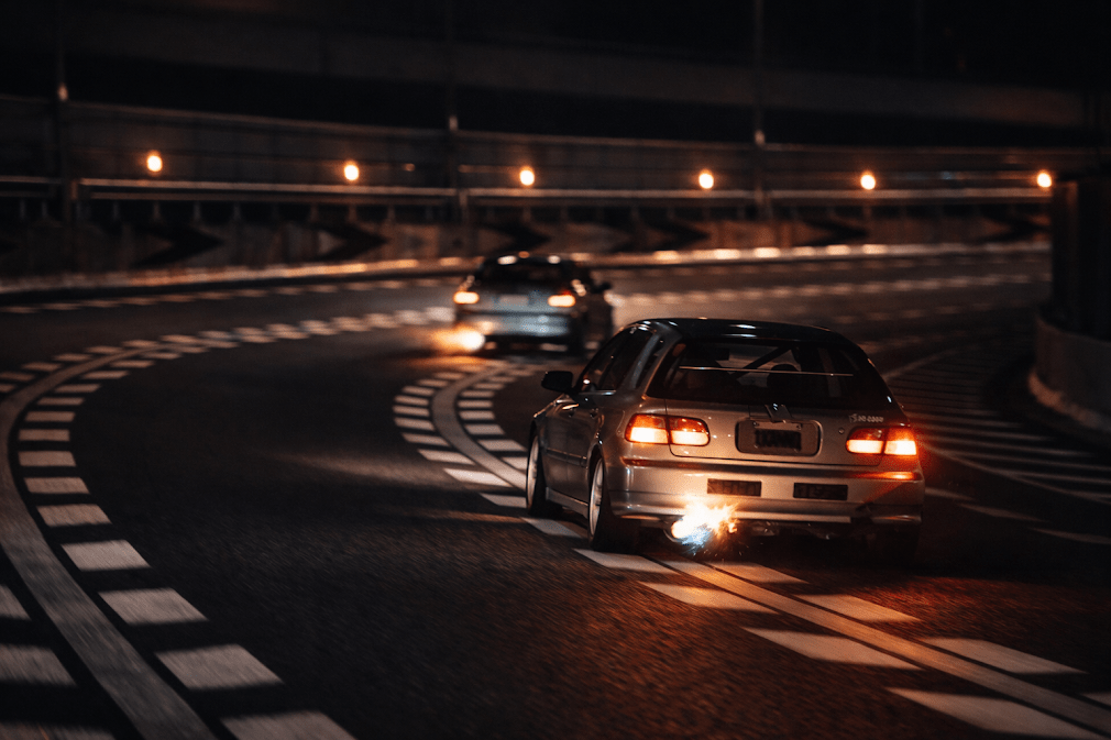 kanjozoku คืออะไร มีที่มาอย่างไร — ภาพรถแฮทช์แบ็กแต่งสไตล์ Honda Civic ขับบนโค้งทางด่วนวงแหวนโอซาก้ายามค่ำคืน มีเปลวไฟจากท่อไอเสีย สะท้อนวัฒนธรรมแก๊งรถซิ่งคันโจโซกุที่เริ่มต้นจากการแข่งบนทางด่วน Loop Line ของญี่ปุ่น