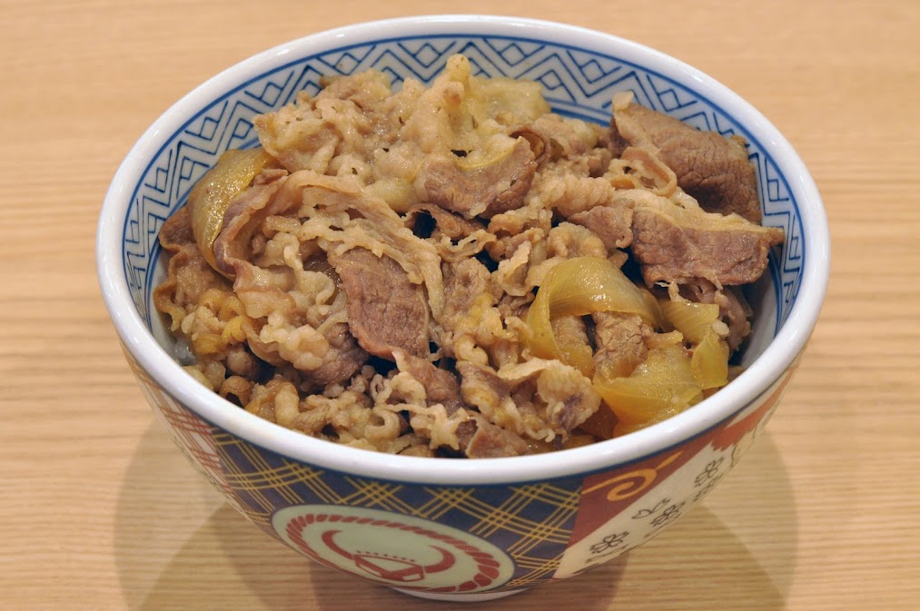 Gemini บอกว่า
ภาพระยะใกล้ของชามข้าวหน้าเนื้อวัว (Gyudon) สไตล์ญี่ปุ่นดั้งเดิม เต็มไปด้วยเนื้อวัวสไลด์บางตุ๋นและหัวหอมผัด ในชามเซรามิกที่มีโลโก้ลายเซ็นที่ ร้านข้าวหน้าเนื้อที่ญี่ปุ่น วางอยู่บนโต๊ะไม้สีอ่อนในร้านอาหาร