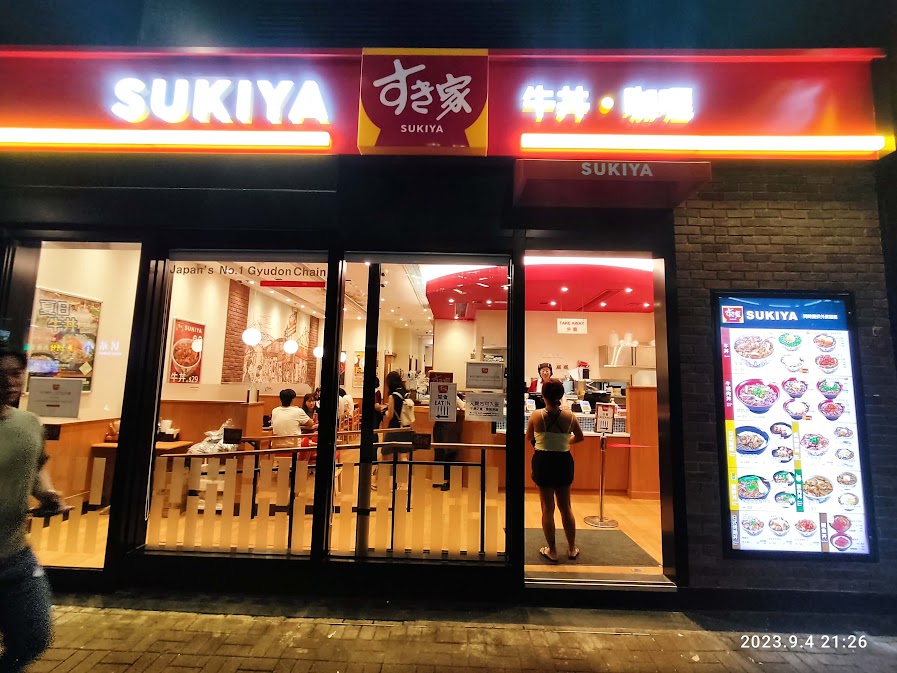 บรรยากาศหน้าร้าน SUKIYA สาขาญี่ปุ่นในยามค่ำคืน ร้านข้าวหน้าเนื้อที่ญี่ปุ่น ยอดนิยม พร้อมป้ายไฟสีแดงสดใส มีเมนูและโลโก้ すき家 ชัดเจน