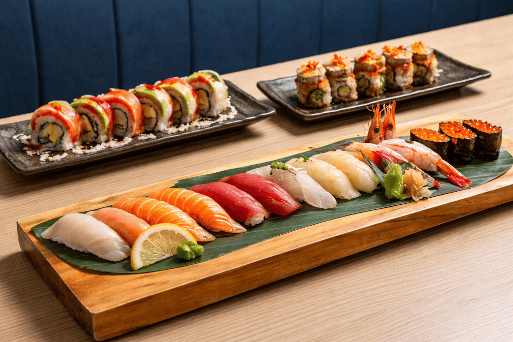 sushi ญี่ปุ่นมีกี่ประเภท ภาพชุดซูชิญี่ปุ่นรวมหลายชนิดบนถาดไม้ มีนิกิริหน้าแซลมอน ทูน่า กุ้ง และไข่ปลา พร้อมโรลซูชิด้านหลัง จัดเสิร์ฟในร้านอาหารสไตล์โมเดิร์น