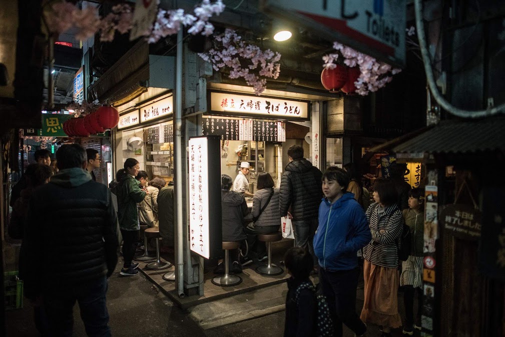 ที่เที่ยว Shinjuku โตเกียว บรรยากาศยามค่ำคืนของร้านอาหารสไตล์ญี่ปุ่นในตรอกเล็กย่านชินจูกุ ผู้คนยืนต่อคิวทานอาหารใต้โคมไฟแดงและดอกซากุระ สะท้อนเสน่ห์สตรีทฟู้ดญี่ปุ่นยอดนิยม