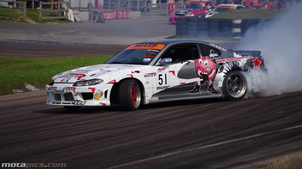 รถ drift ญี่ปุ่น Nissan Silvia S15 กำลังดริฟต์ในสนามแข่ง มีควันยางลอยด้านหลัง พร้อมลวดลายแต่งรถสไตล์ JDM แสดงเอกลักษณ์รถดริฟต์ญี่ปุ่นระดับมืออาชีพ 🚗💨🇯🇵