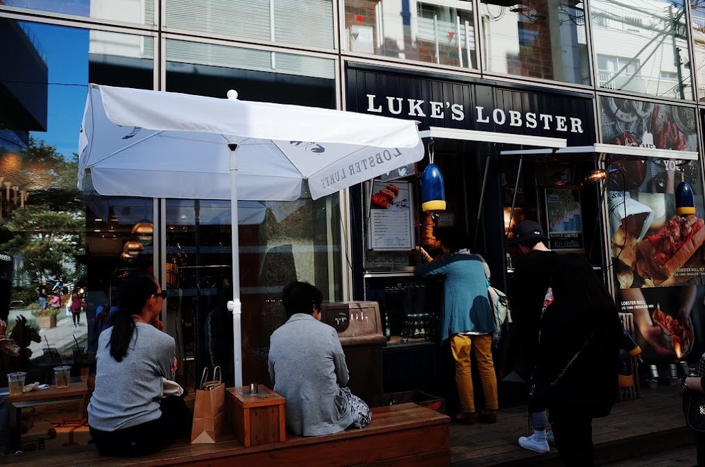 บรรยากาศหน้าร้าน Luke's Lobster ร้านแซนด์วิชล็อบสเตอร์โรลชื่อดังที่เป็นหนึ่งในของกินย่าน Harajuku ยอดนิยม มีลูกค้ากำลังยืนต่อคิวสั่งอาหารและนั่งรอหน้าร้านตกแต่งสไตล์โมเดิร์นคลาสสิก
