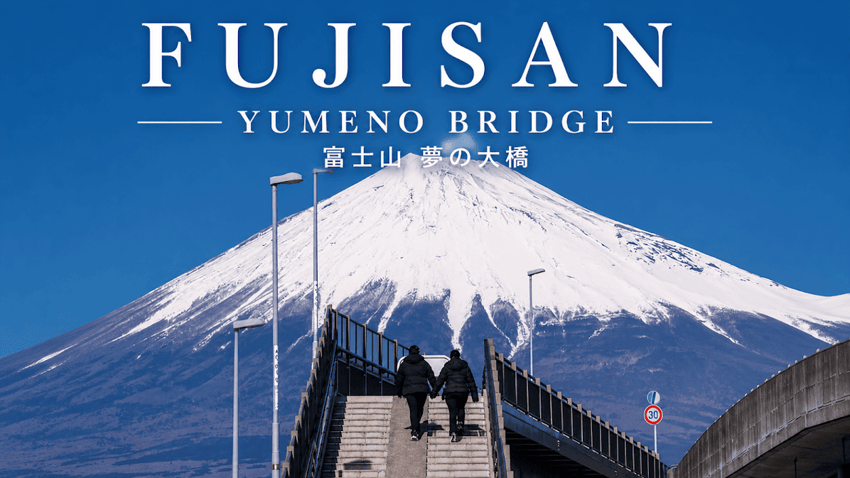 ภาพสะพาน Yumeno Bridge ในญี่ปุ่นที่มีผู้คนกำลังเดินขึ้นบันได โดยมีภูเขาไฟฟูจิที่ปกคลุมด้วยหิมะขนาดใหญ่โดดเด่นอยู่ด้านหลัง ท้องฟ้าสีฟ้าสดใสและบรรยากาศฤดูหนาวช่วยเพิ่มความสวยงามของวิว จุดนี้เป็นหนึ่งในมุมถ่ายภาพยอดนิยมสำหรับคอนเทนต์ “10 จุดถ่ายรูปภูเขาไฟฟูจิ 2026”