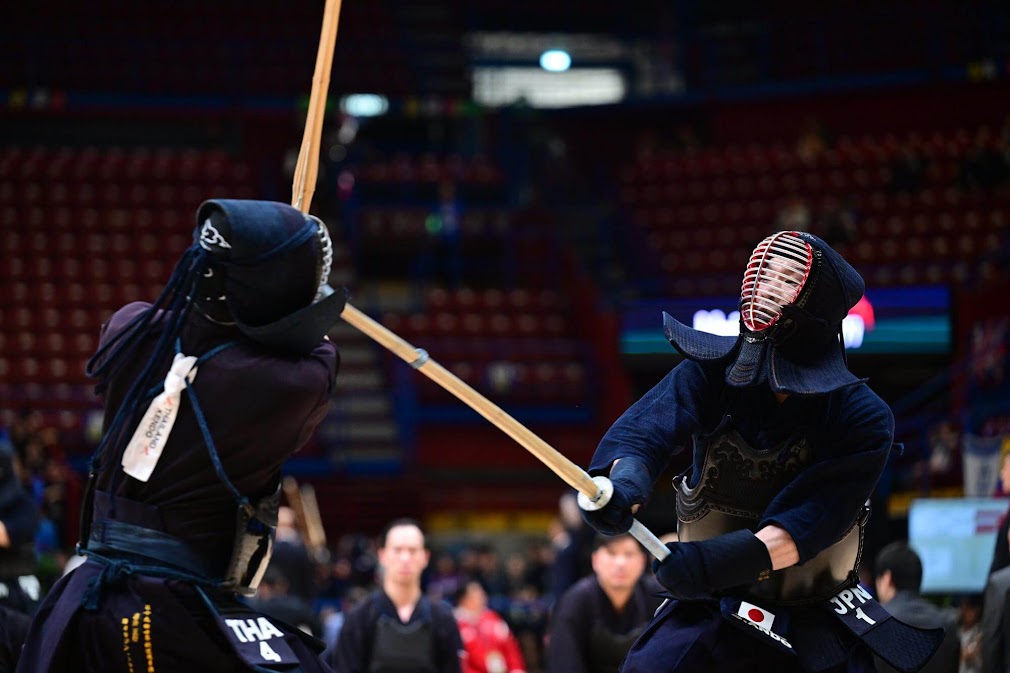 นักกีฬาสองคนกำลังประลองฟันดาบเคนโด (kendo) อย่างเข้มข้นในสนามแข่งขัน สวมชุดเกราะและหน้ากากแบบดั้งเดิม ใช้ดาบไม้ไผ่ปะทะกันกลางอากาศ โดยมีผู้ชมและกรรมการอยู่ด้านหลัง แสดงให้เห็นว่า “ฟันดาบ kendo คืออะไร” ผ่านการต่อสู้ที่เน้นความเร็ว ความแม่นยำ และวินัยของศิลปะการต่อสู้ญี่ปุ่น