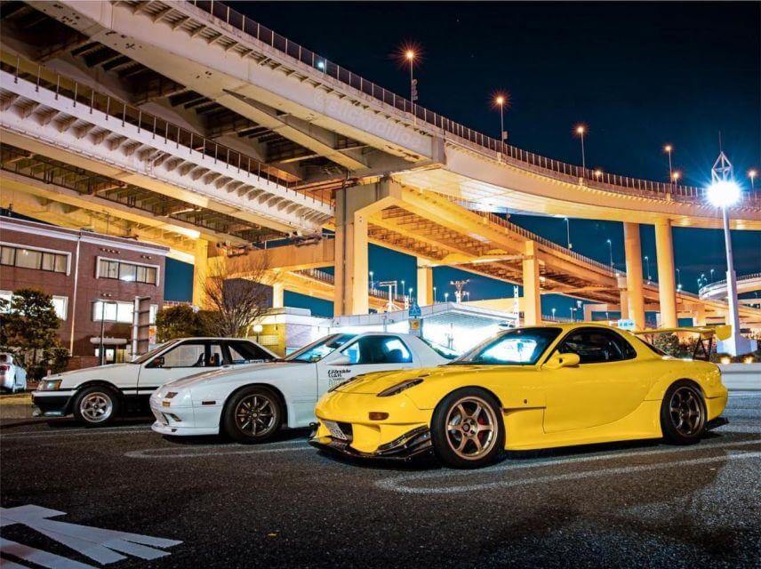 ภาพถ่ายยามค่ำคืนของกลุ่มรถยนต์ สไตล์แต่งรถญี่ปุ่น JDM (เช่น Toyota AE86, Mazda RX-7) ที่จอดเรียงกันที่ Daikoku PA (PA 73) ในโตเกียว ประเทศญี่ปุ่น พร้อมคำบรรยายภาษาไทยระบุชิ้นส่วนที่ได้รับการปรับแต่ง เช่น ชุดแต่งตัวถังแบบ Widebody และเครื่องยนต์ Rotary ภายใต้แสงไฟสีทองของทางยกระดับที่เป็นสัญลักษณ์ของย่านอุตสาหกรรม"