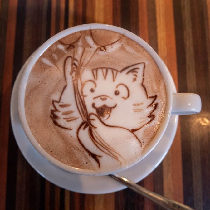ภาพมุมสูงของถ้วยกาแฟเซรามิกสีขาวบนจานรอง เผยให้เห็นฟองนมลาเต้อาร์ตเป็นรูปวาดลายเส้นแมวลายเสือกำลังยิ้มและถือช่อลูกโป่ง ของกินย่าน Harajuku วางอยู่บนโต๊ะไม้ที่มีลวดลายทางยาวสีน้ำตาลสลับเข้มอ่อน