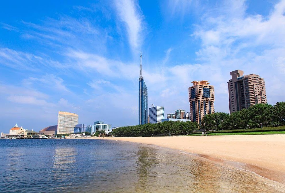 ชายหาดโมโมจิในฟุกุโอกะ น้ำทะเลใส ทรายสีทอง พร้อมวิวเมืองและตึกสูง เช่น Fukuoka Tower ภายใต้ท้องฟ้าสีฟ้า เหมาะสำหรับแวะพักผ่อนระหว่าง ขับรถเที่ยวฟุกุโอกะ รถเช่าญี่ปุ่น