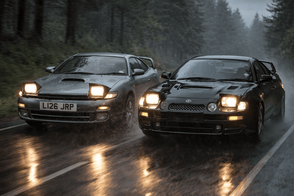 Toyota Celica สองคันสีเทาและสีดำขับเคียงกันบนถนนเปียกกลางป่าสนในวันที่ฝนตก ไฟหน้าเปิดสะท้อนบนพื้นถนน สื่ออารมณ์สปอร์ตดุดันและพลังการขับขี่ของ toyota celica
