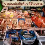 ภาพหน้าแผงขายอาหารทะเลสดที่เต็มไปด้วยปลา ปูยักษ์ และหอยนานาชนิดใน ตลาดปลา ใน ญี่ปุ่น พร้อมป้ายราคาภาษาญี่ปุ่นปักอยู่ตามถาดน้ำแข็ง โดยที่ด้านบนของภาพมีกล่องข้อความพาดหัวสีน้ำตาลสไตล์พู่กันเขียนว่า "5 ตลาดปลาโตเกียว ที่ห้ามพลาด" เพื่อใช้เป็นภาพหน้าปกหรือภาพประกอบบทความท่องเที่ยว ด้านหลังมีพ่อค้ากำลังยืนให้บริการลูกค้าในบรรยากาศตลาดสดแบบดั้งเดิม